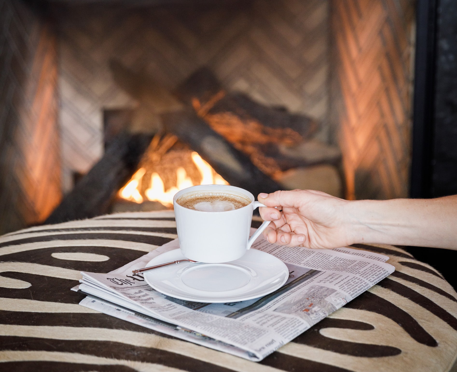 Woman reaching for a cappucino in front of a fireplace.